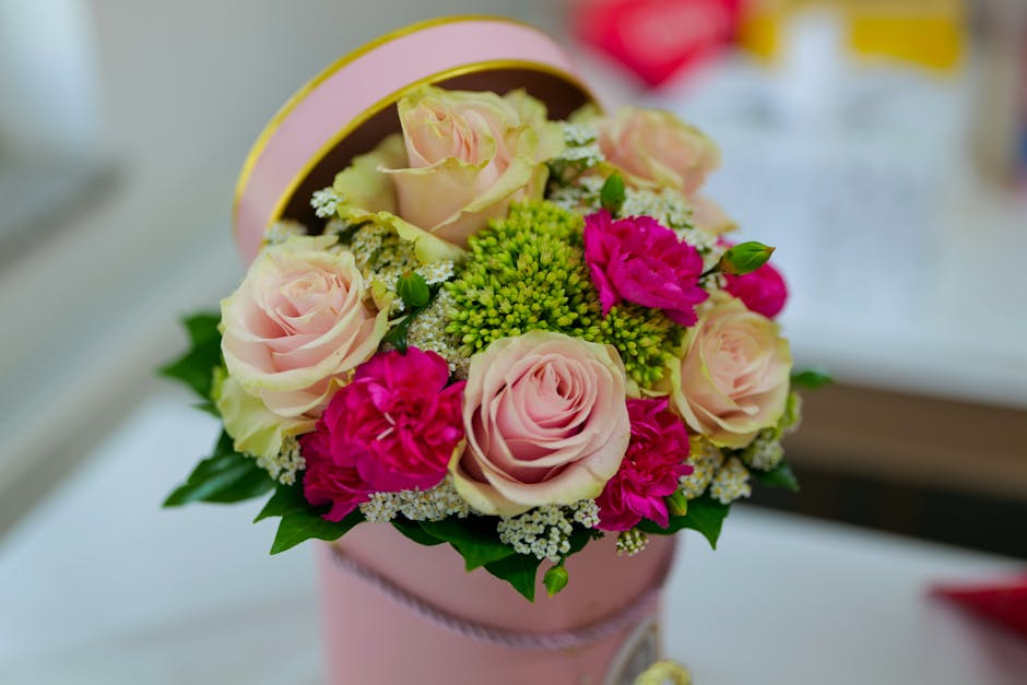 A round, pastel pink floral gift box filled with a fresh bouquet of pale pink roses, vibrant fuchsia carnations, and small white fillers, arranged with sprigs of green foliage. The flowers are neatly
