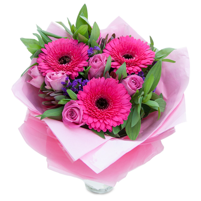 I see this bouquet as a study in pinks and textures: three confident fuchsia gerberas dominate the scene, their broad, sunburst petals arranged with precision around deep, almost-black centres that create a striking focal point. Nearby, roses in a dusty lavender-pink soften the overall mood with layered, velvety petals that open like little cups of scent-gentle, powdery perfume rather than overt sweetness. Tiny clusters of purple statice are thoughtfully tucked between the larger blooms, their papery, compact florets bringing a tactile counterpoint and a cool violet note against the warmer roses and gerberas. Lush greenery-leaves that read as salal or eucalyptus-wraps the blooms in a verdant frame, offering both visual weight and a hint of crisp, green aroma. The bouquet is swathed in an inner blanket of soft pale pink tissue; over that, a slightly crinkled translucent cellophane in a vibrant fuchsia-pink gives the whole piece a subtle glow and a touch of glamour without feeling fussy. The lower portion reveals the top rim of a clear glass vase, so you can imagine placing it immediately on a dining table or office desk. Bathed in soft daylight, the colours hold a luminous clarity that lifts the spirit-this is the sort of arrangement that feels equally at home on a birthday table or as a thoughtful surprise delivered to a neighbour near Becontree or Dagenham Dock.